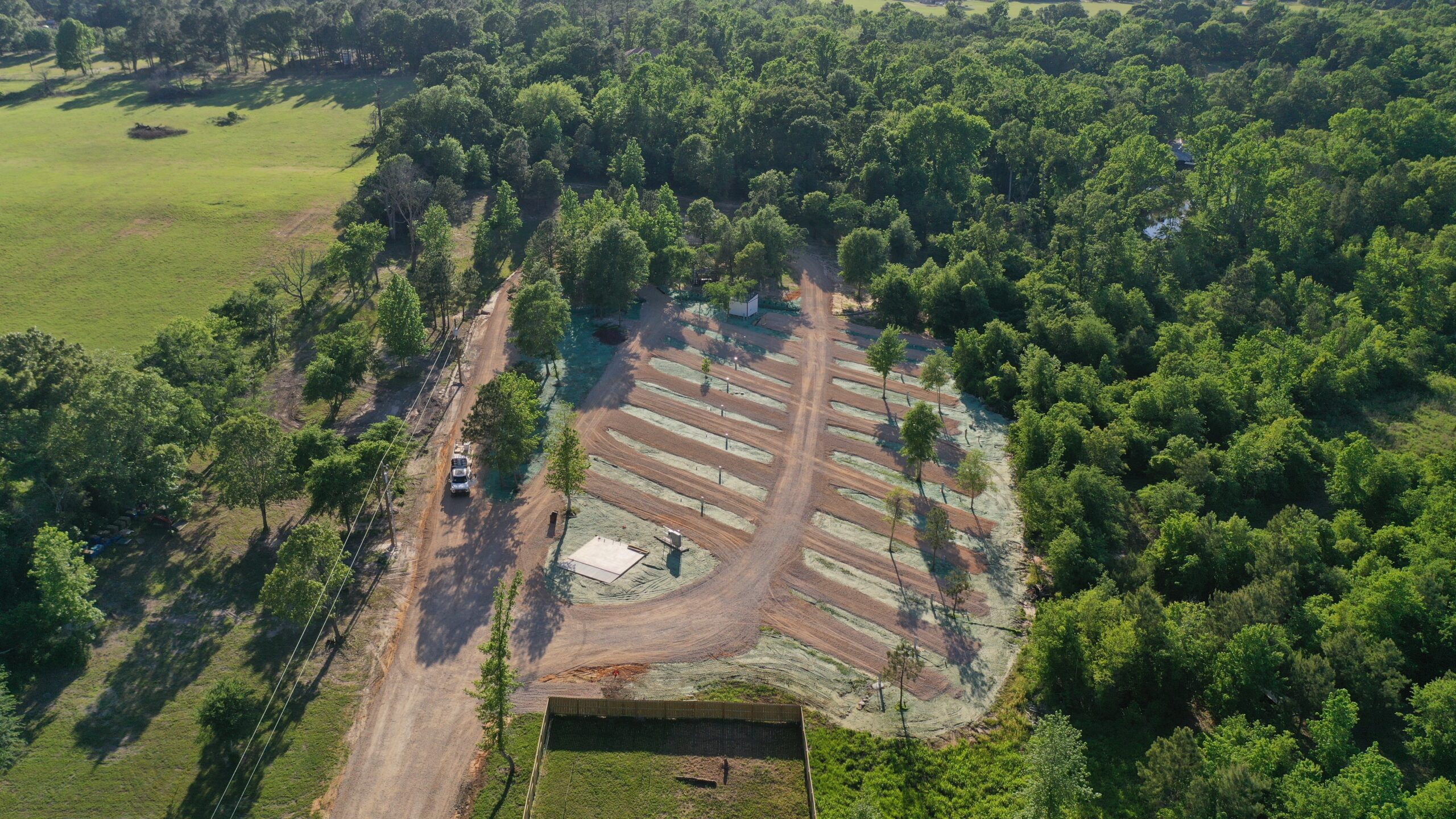 Aerial view of a newly developed RV park in a forested area, showing empty gravel parking spots lined with trees, dirt roads, and a few parked vehicles. Surrounding areas are densely wooded with open grassy fields nearby.