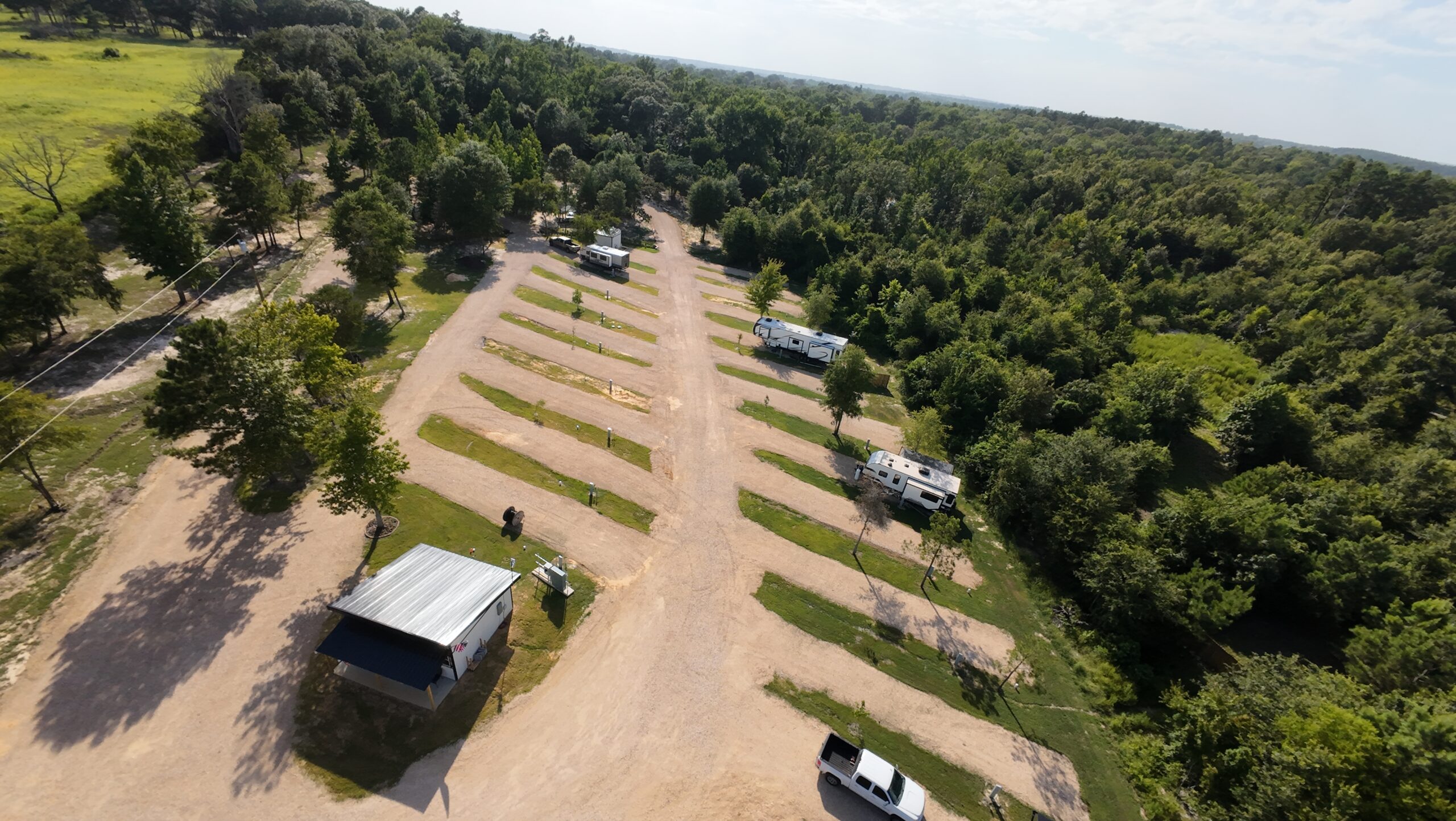 Aerial view of an RV campground with several empty gravel parking spaces, a few parked RVs, a small building, and surrounding green trees under a partly cloudy sky.