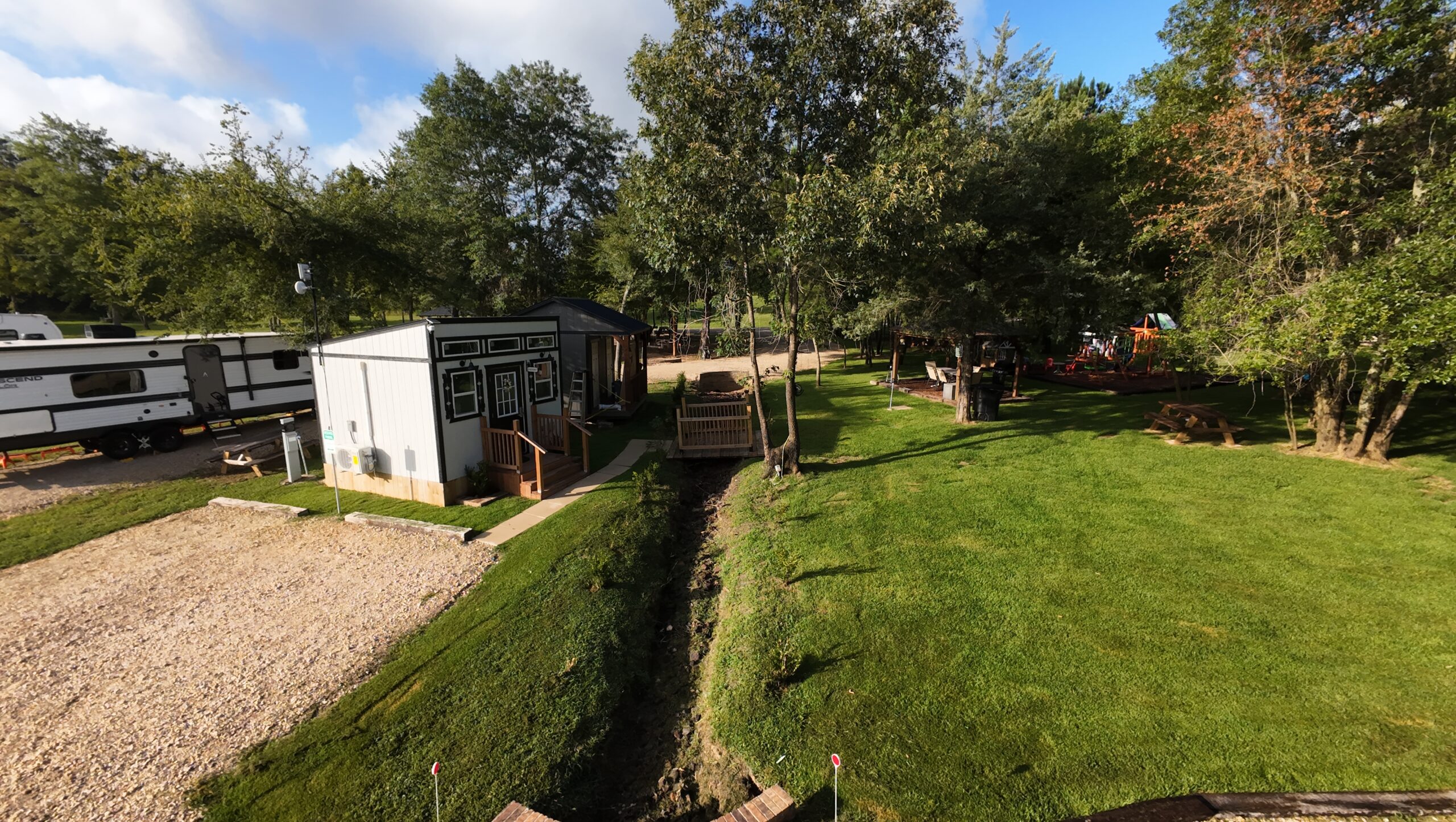 Small house and RV parked on gravel near a grassy yard with trees, a wooden bridge over a creek, and picnic tables under the trees on a sunny day.